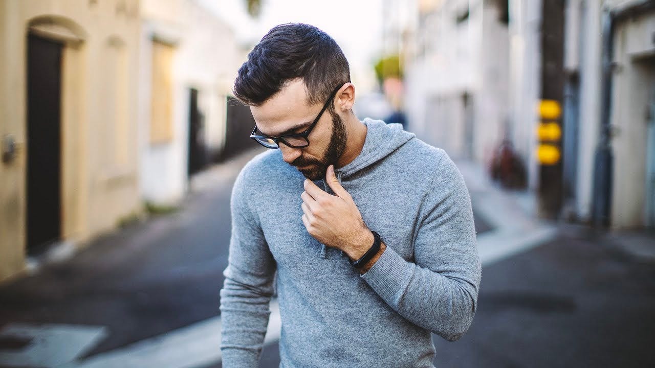 YouTuber Matt D’Avella looking right and down with his left hand on his thin. He’s wearing glasses, a black watch on his left wrist and a long sleeve gray shirt with a hood. He’s standing in the middle of the street, but it is blurred.