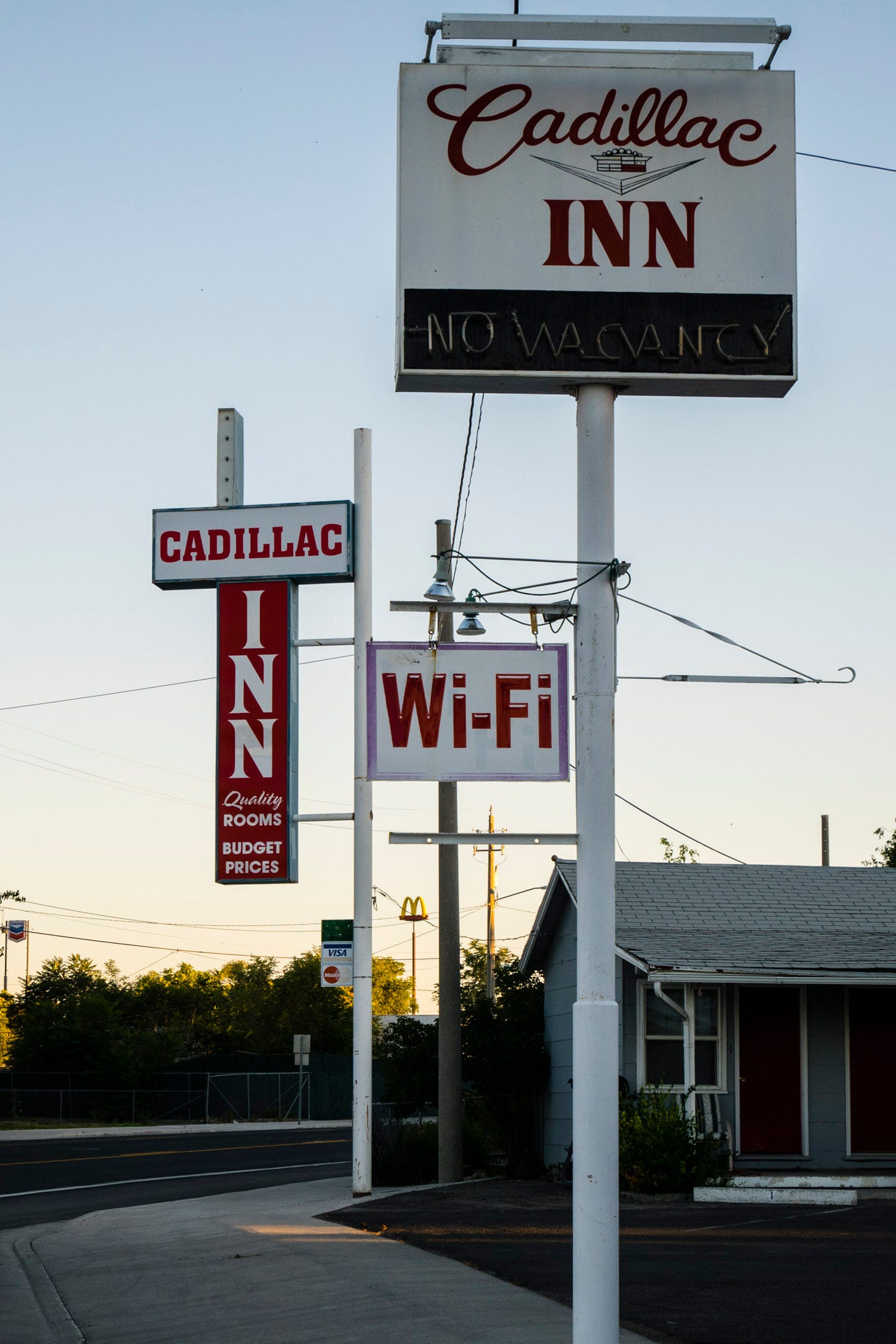 Photo of roadside signs on metal poles painted white at the entrance to a motel. The closest sign is white with "Cadillac Inn" in a dark red. Cadillac is written in decorative script, Inn is all caps, serif, between the words is the crest of Cadillac automobiles. The is a light fixture on top of the sign to illuminate the name. Below is a black painted area with an unlit neon "NO VACANCY" sign. Further down and hanging off the side of the pole is a white "WiFi" sign with inactive spotlights pointed at it. On the other side is another horizontal protuberance with a hook on the end. Nothing is hanging from it.  O the other side of the motel driveway is another Cadillac Inn sign on another white pole. This one looks newer and is internally lit. It has CADILLAC in all caps, block serif, red letters on white on top. Hanging below is a vertical red sign with white letters, with INN written vertically, below which is written "Quality (italics) ROOMS" on two lines, and below that "BUDGET PRICES" on two lines. In the right of the photo is the corner of the motel. Beyond are some tall bushes and beyond that are tall signs for McDonalds and Chevron on very large poles.