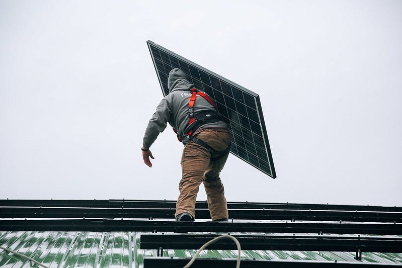 A worker on a roof carries a solar panel on one shoulder as part of a rooftop solar installation project.