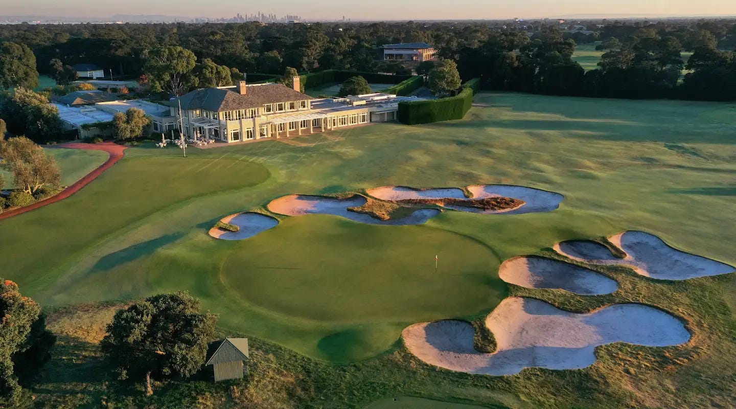 Aerial view of a luxurious golf course with a large clubhouse, multiple water hazards, and sand bunkers surrounded by lush greenery and trees. A city skyline is visible in the distance.