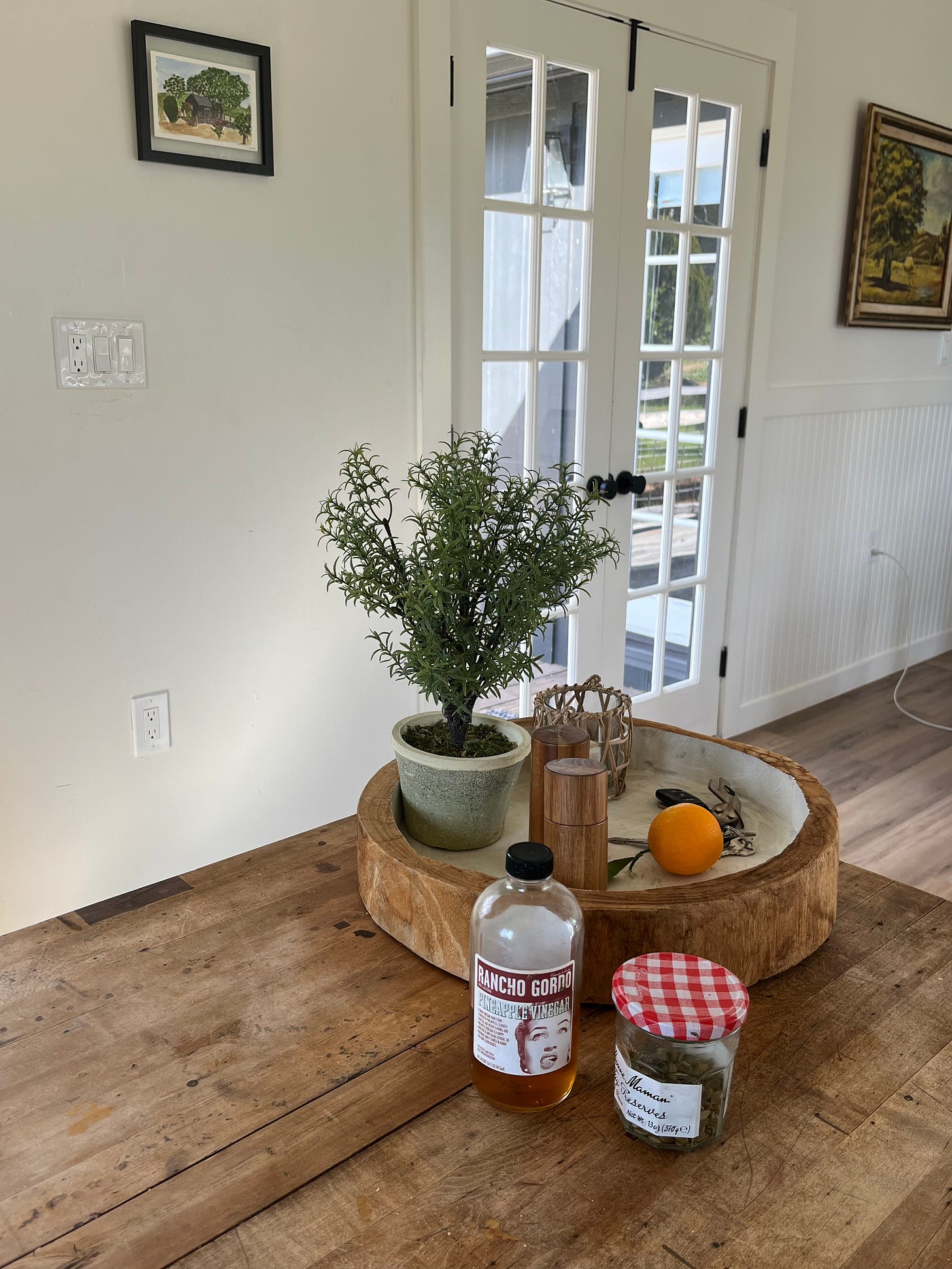 A farmhouse table with vinegar, jar of pumpkin seeds, and a small olive tree.