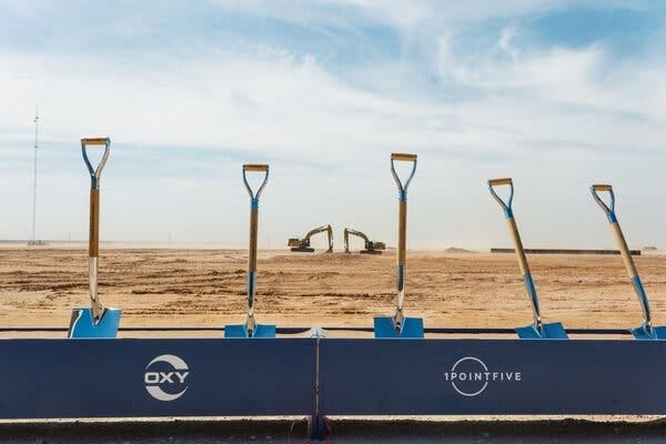 Five brand-new blue shovels are lined up in the brown dirt of an empty field, ready for a groundbreaking ceremony. 