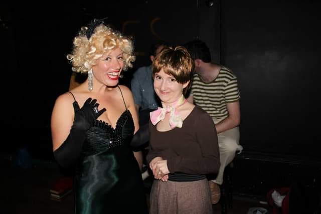 A woman in a blonde wig dressed as Marilyn Monroe stands next to a woman in a dowdy brown top and skirt with a short brown wig. Both are smiling