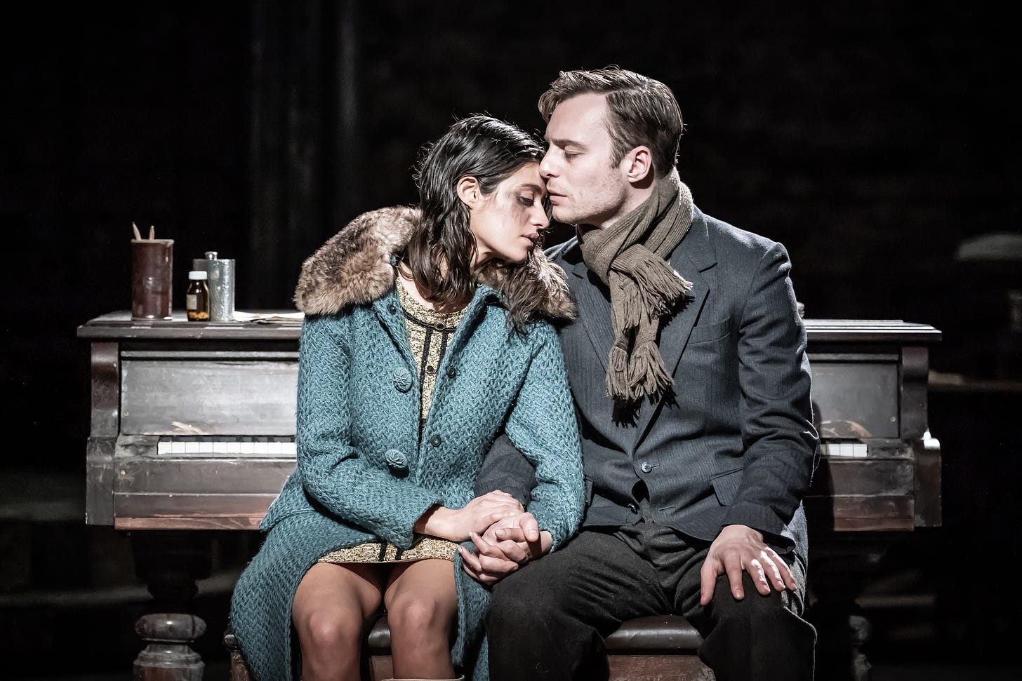 A woman and a man sit on a piano stool, holding hands. They look tired and cold.