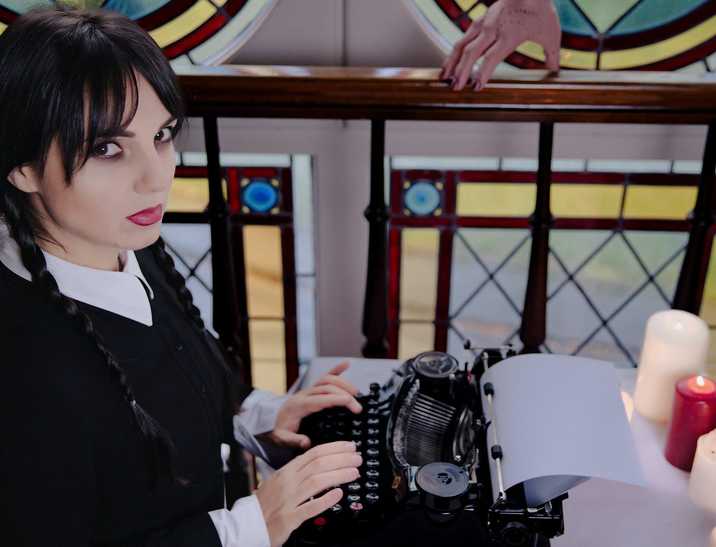 photo entitled "Girl in the Wednesday Addams Costume Writing on a Typewriter." a goth-looking young woman looks at the camera while her hands are positioned on the keys of a typewriter. the photo appears to be taken in a church, as there are stained glass windows behind her. candles are lit on the table upon which the typewriter sits. a disembodied hand sits on a bannister in the background.