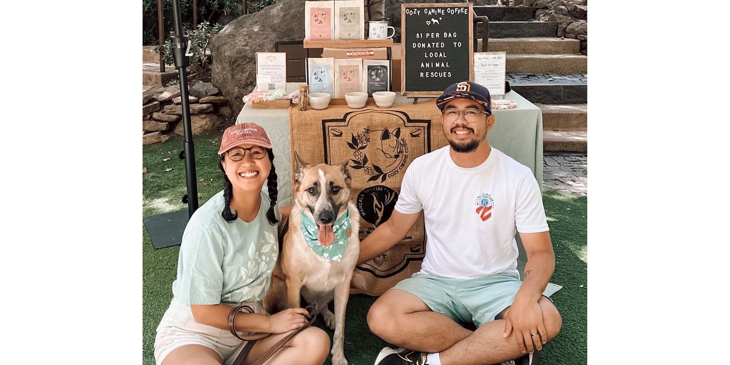 A couple sits cross-legged on the grass. Their dog sits tall between them. They smile for the camera posed in front of a table with coffee bags and merchandise at a local outdoor marketplace.