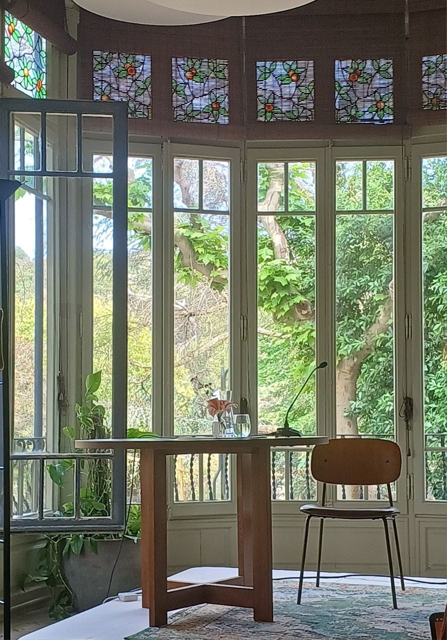 Fotografia di un ambiente interno molto luminoso, con una vetrata ad arco composta da pannelli di vetro decorati in alto con motivi floreali colorati. Davanti alla vetrata c’è un tavolino in legno con un microfono, un bicchiere d’acqua, una brocca e un piccolo vaso con fiori. Accanto al tavolo, una sedia in legno chiaro con struttura in metallo. Ai piedi del tavolo, un tappeto con motivi orientali. Sullo sfondo, si vedono alberi e vegetazione attraverso i vetri. Fotografia di un ambiente interno molto luminoso, con una vetrata ad arco composta da pannelli di vetro decorati in alto con motivi floreali colorati. Davanti alla vetrata c’è un tavolino in legno con un microfono, un bicchiere d’acqua, una brocca e un piccolo vaso con fiori. Accanto al tavolo, una sedia in legno chiaro con struttura in metallo. Ai piedi del tavolo, un tappeto con motivi orientali. Sullo sfondo, si vedono alberi e vegetazione attraverso i vetri.