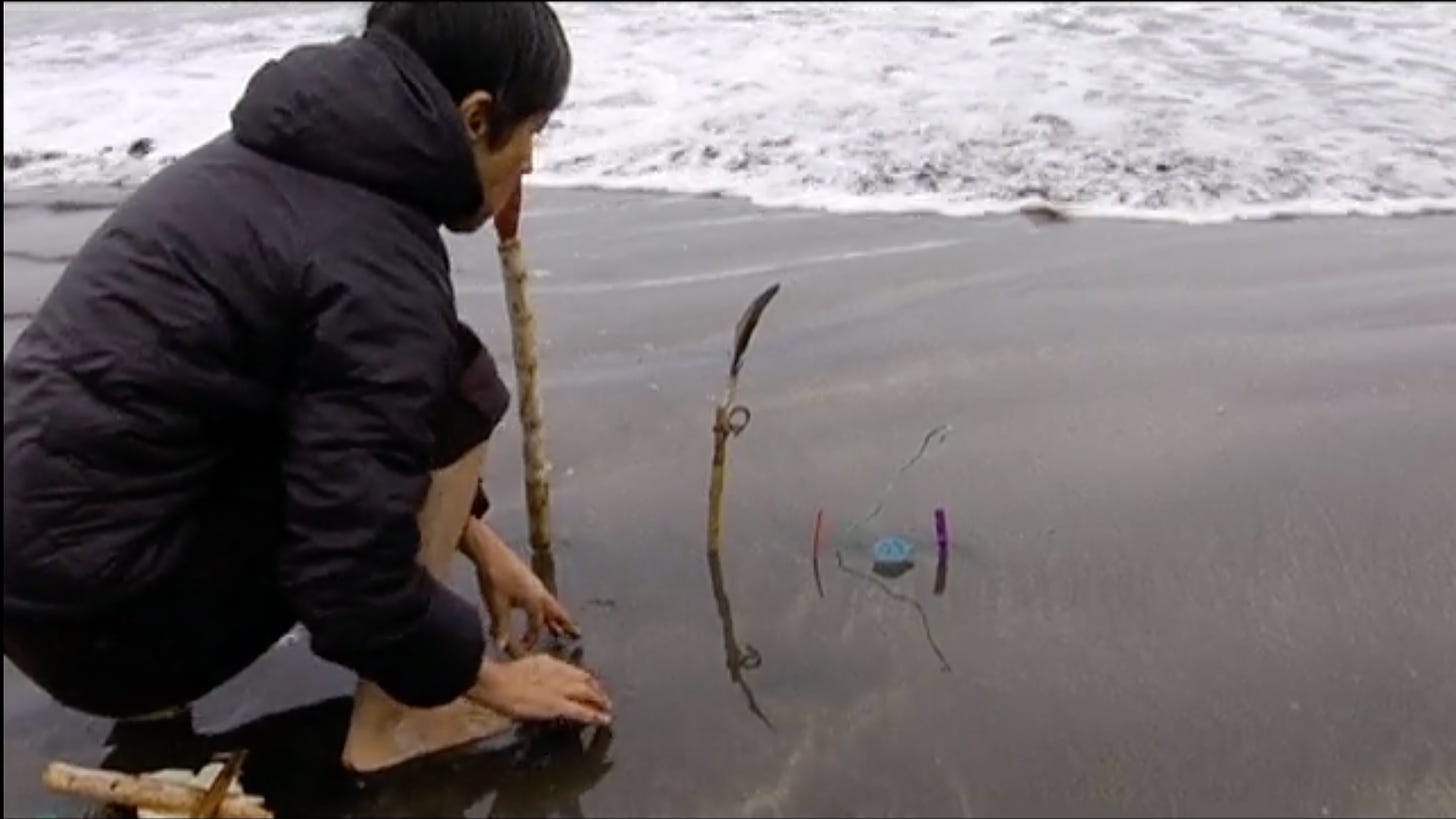 A woman with short dark hair, wearing a black puffy jacket and bare feet, kneels on the sand in front of a shallow frothy wave. She is setting fragile objects made from beach debris into the sand in the path of the oncoming water.