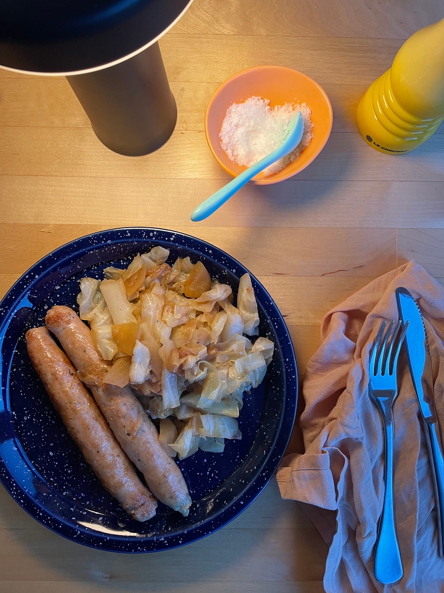 Chicken sausages on a speckled plate alongside braised cabbage, apple and onion. Sea salt and a pepper grinder nearby.