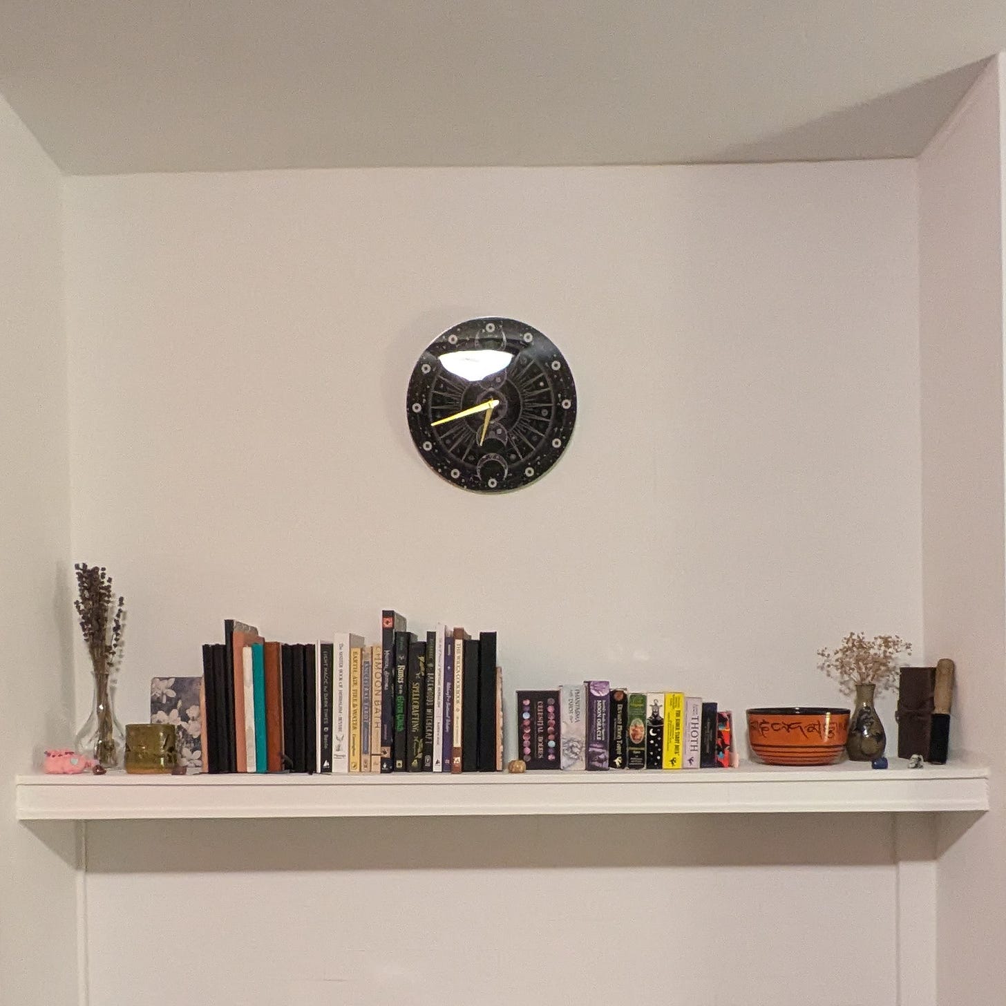 A white mantle with lines of books, tarot cards, a singing bowl, vases of dried flowers, and stones. Above the mantle is a moon-themed analogue clock with the time 6:43.