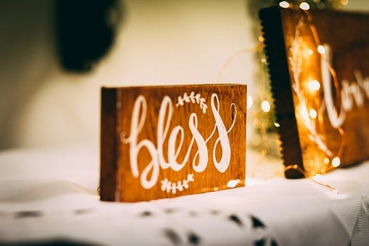 The word “bless”, painted in white on a brown wooden block, placed on a white tablecloth.