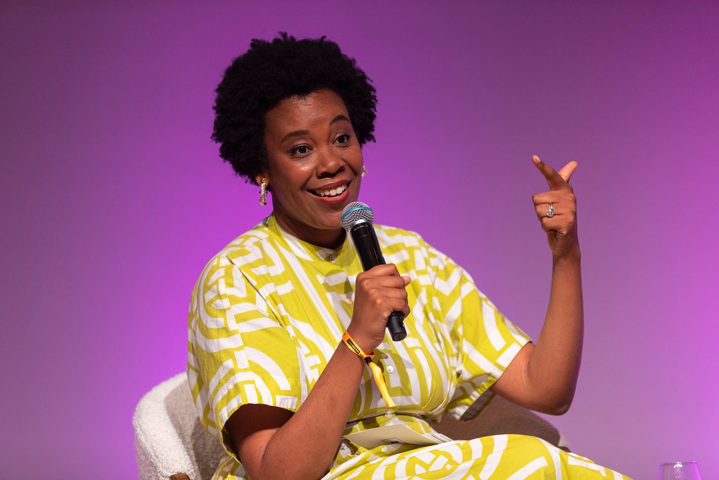 Jen seated and holding a microphone, wearing a patterned yellow dress and with a purple background behind her