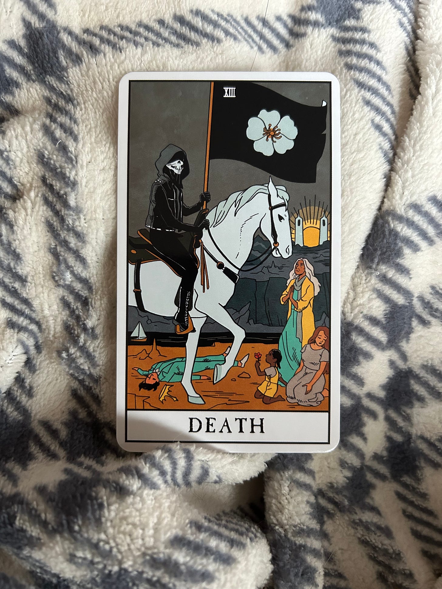 image of the Death card from The Modern Witch Tarot. a cloaked figure with the face of a skeleton sits atop a white horse, holding a black flag with a fllower on it. people in front of the figure and their horse appear to be pleading with them, a small child is on their knees holding a flower out to the figure. beneath the horse lies a figure in a suit with a crown to their side, presumably a king.