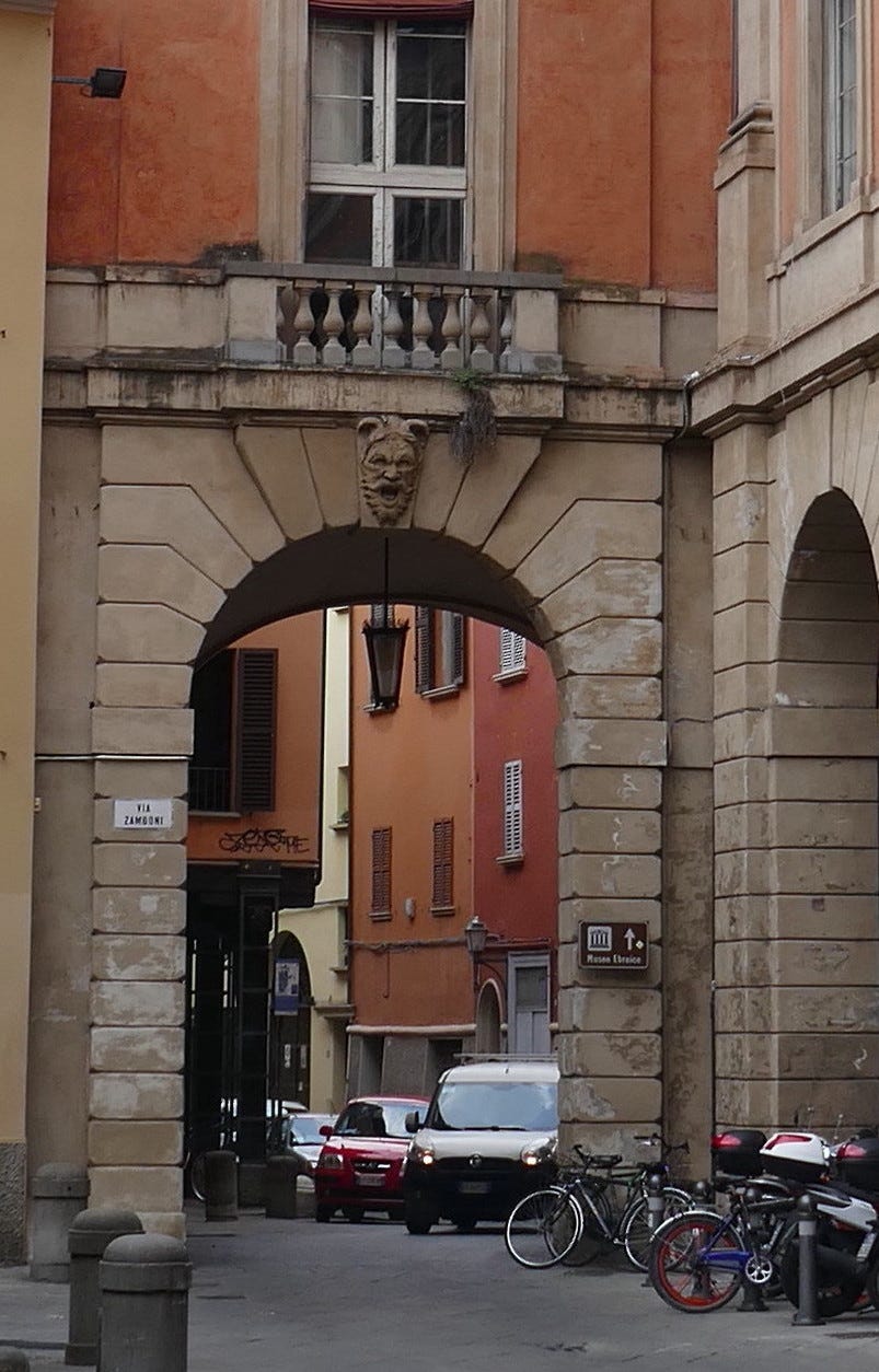 An archway in Bologna with a gargoyle's face overhead and red and orange buildings