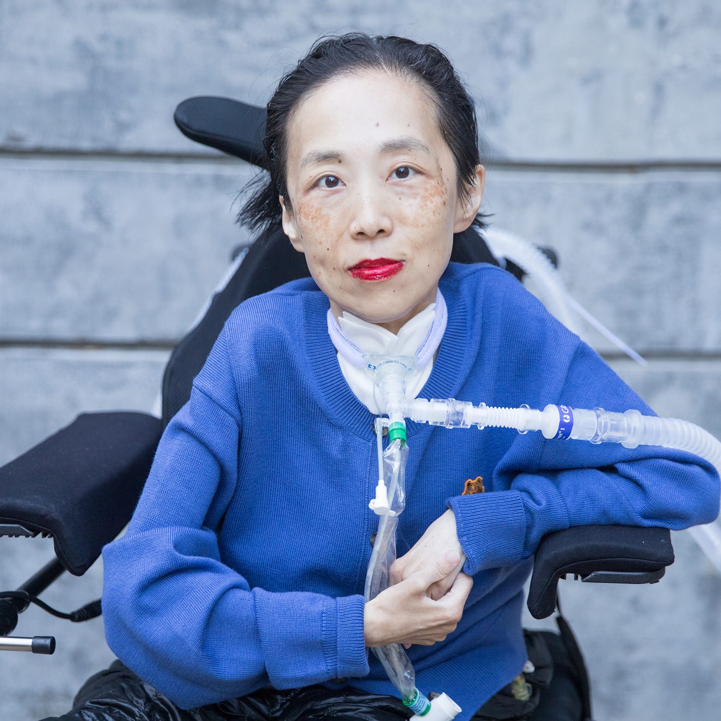 Photo of Alice Wong in front of a cement wall. She is wearing a blue cardigan and sitting in a power chair. She is staring intently at the camera, wearing a bold red lip color, and a trach at her neck. Photo credit: Eddie Hernandez Photography.