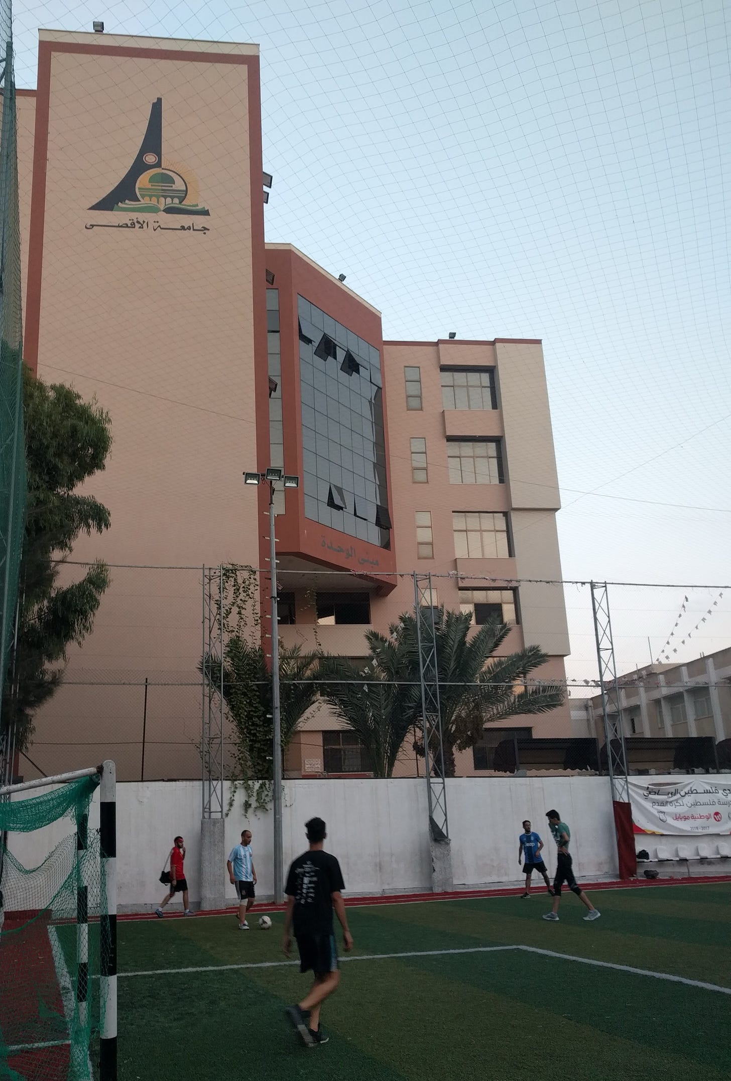 Men play soccer on a fenced-in turf field with a large building behind them