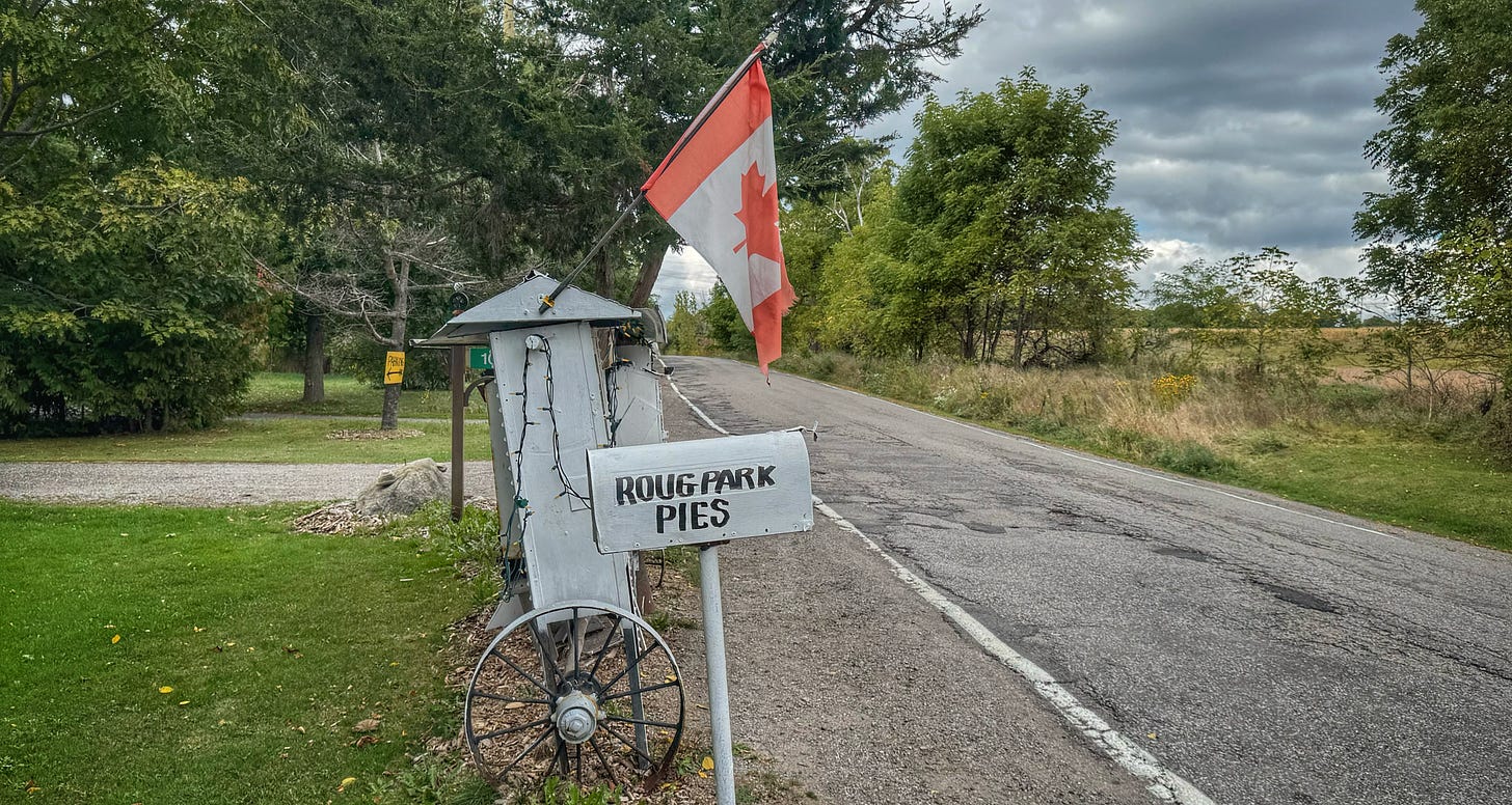 A side view of the Rouge Park Pie Stand, taken from the side of the road. In the foreground is a weathered white mail box that reads "Roug Park Pies." There is a Canadian flag attached to the roof of the stand and a steel wagon wheel attached to its legs. 