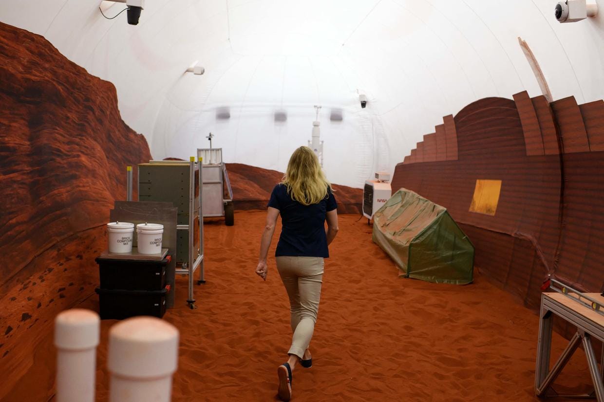 A woman walks across a room with a floor of red sand and a white domed ceiling.