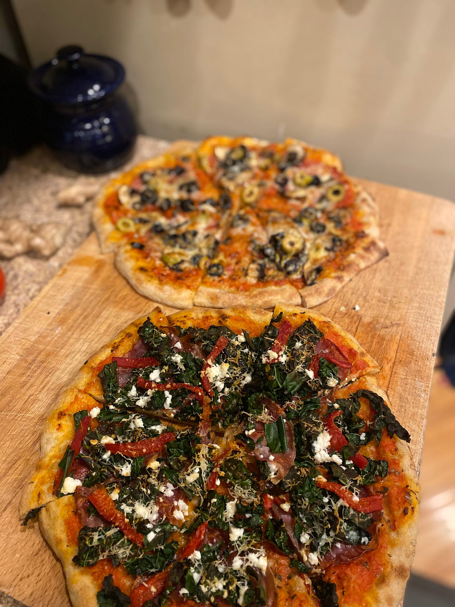 The pizzas described above, sliced into eighths on a cutting board. The kale pizza is in the foreground, the olive a little blurred in the back.