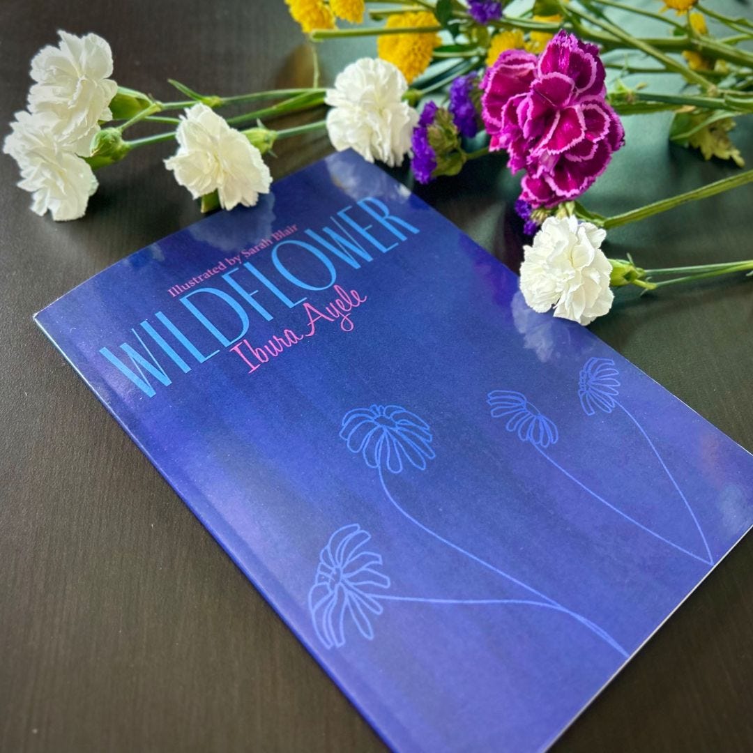 Photo of Wildflower book. Cover is purple with light blue and pink writing. It reads: Illustrated by Sarah Blair, Wildflower Ibura Ayele. The book sits on a dark wooden table next to pink, white, and 