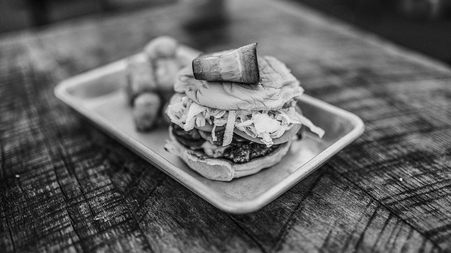 A burger sits on a metal tray on top of a raw wood table