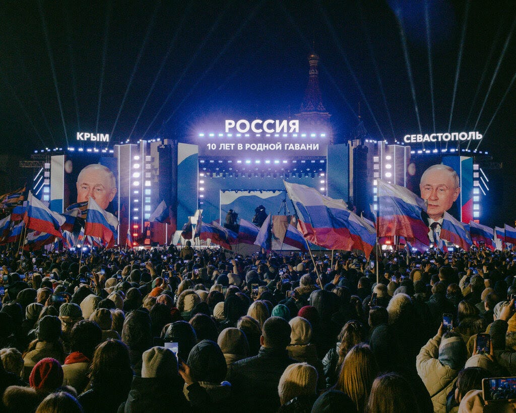 People with Russian flags gathering in front of a stage with two giant screens showing Vladimir Putin.
