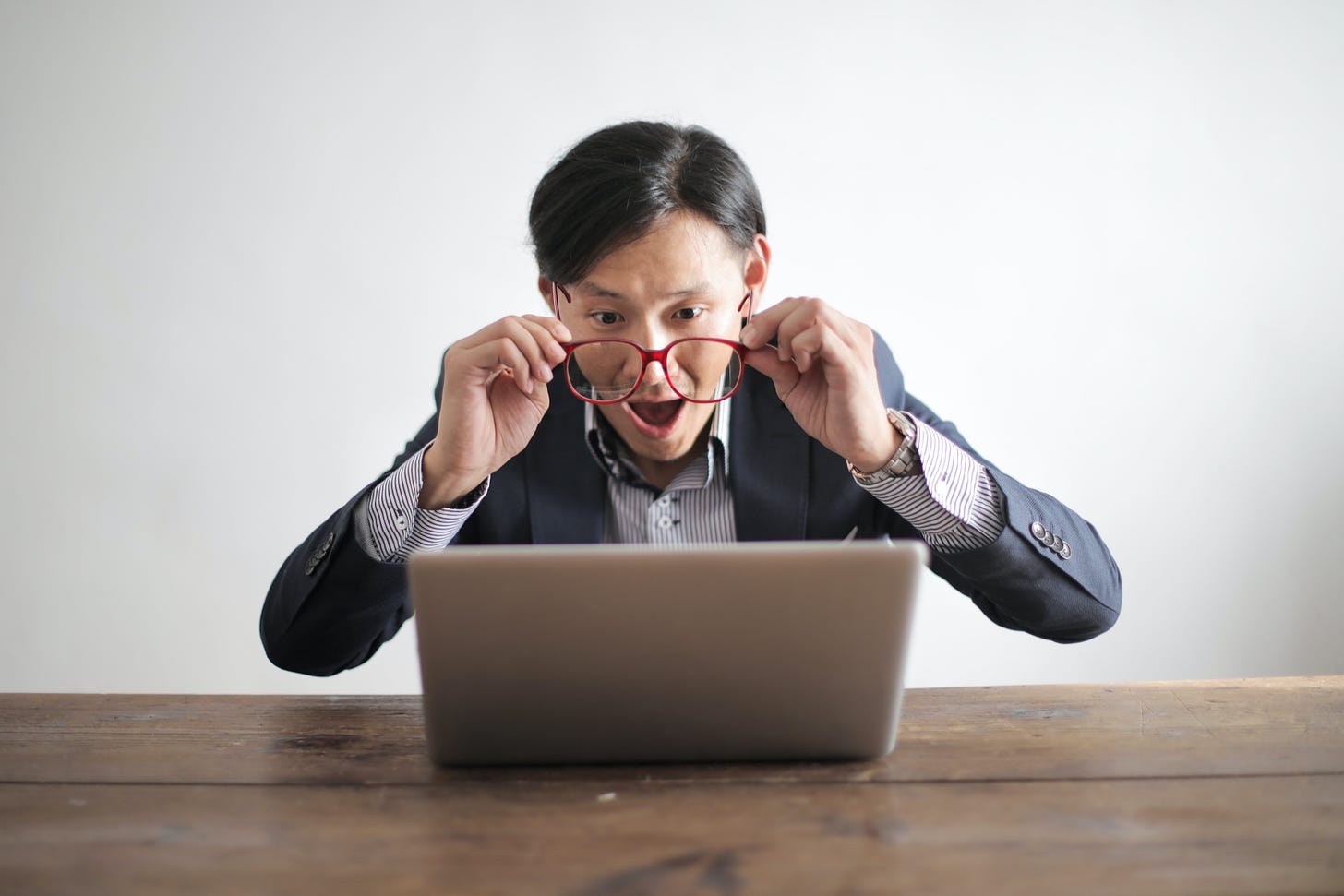 Amazed male in business casual pulling glasses off in front of laptop on table.