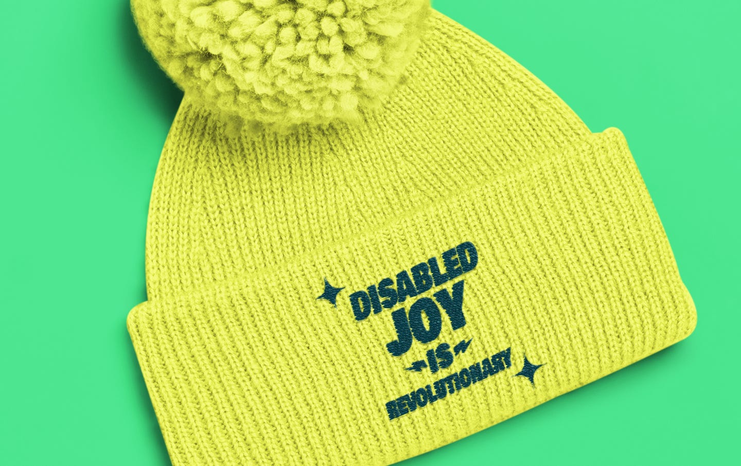 This vibrant yellow pom-pom beanie, known as the Disability Joy Knitted Pom Pom Hat by Disability Culture Lab, features a prominent folded brim adorned with "Disabled Joy is Revolutionary" in bold black letters against a striking green background. This design highlights the exceptional union-embroidered craftsmanship of renowned disabled artist Jennifer White-Johnson.