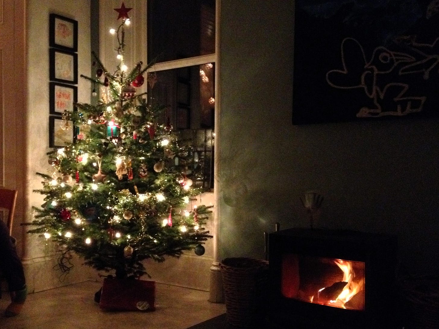 A lit up christmas tree standing in the corner of a room lit also by a stove