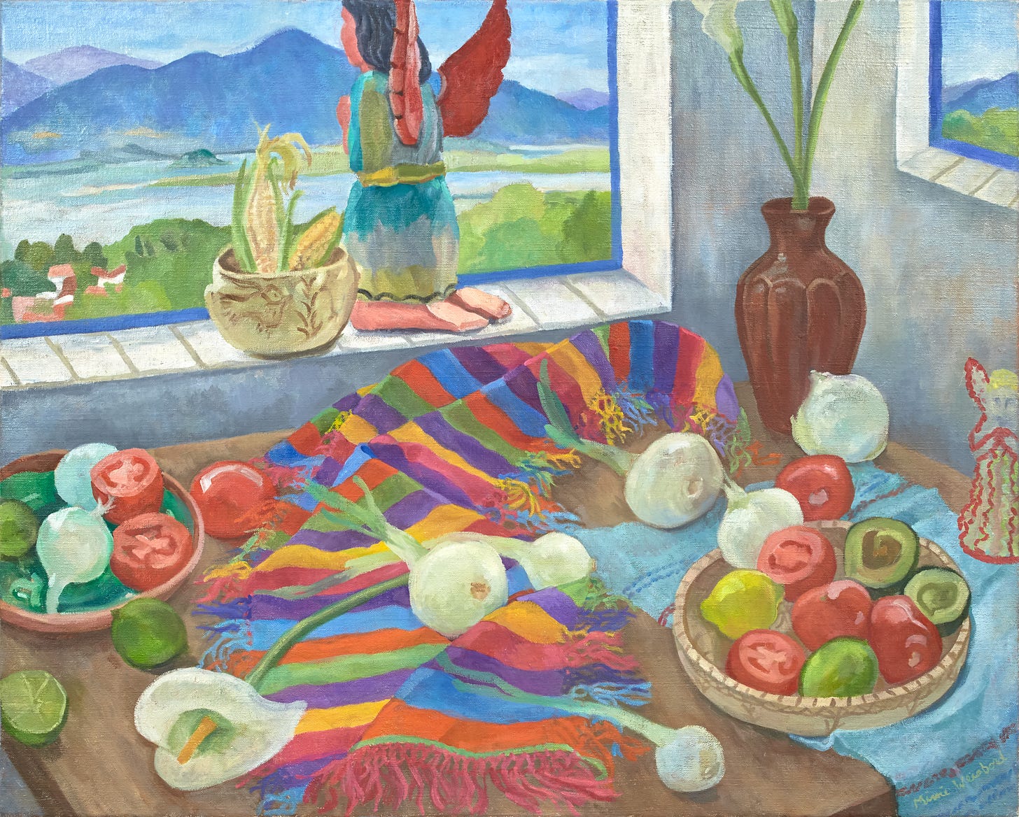 A table holds a Mexican cloth in rainbow colors beneath a calla lily and sprouting onion bulbs. The table also has bowls of cut and whole tomatoes, avocados, a vase in the corner, a small angel figurine on the side, and on the window ledge behind is a painted bowl with ears of corn and a large kneeling angel figure in a prayer position facing the landscape of mountains, lake, and a town nestled in the trees below.