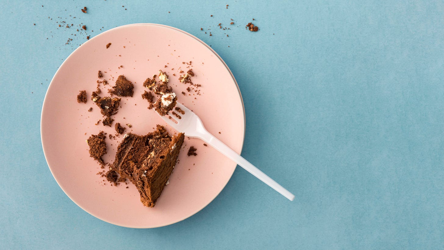 A piece of chocolate cake, mainly crumbs, on a pink plate. The background is pale blue. 