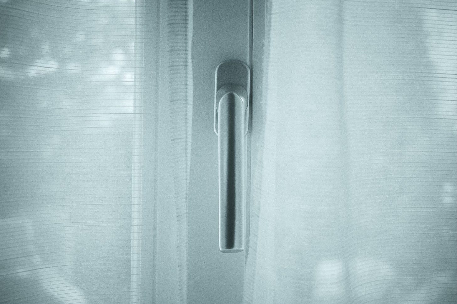 A close-up of a modern window handle centered between two sheer white curtains, with soft natural light filtering through the fabric. The overall image has a cool, desaturated tone.
