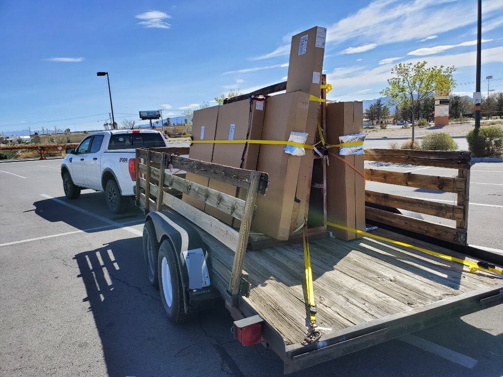 A white truck pulling a trailer loaded with tall cardboard boxes on a window rack.