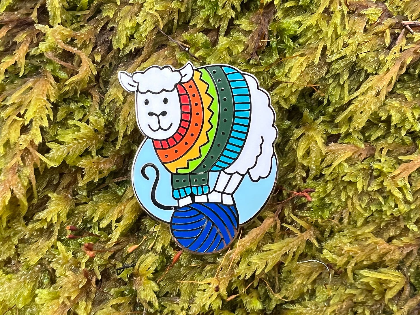 An enamel pin of a sheep wearing a rainbow jumper