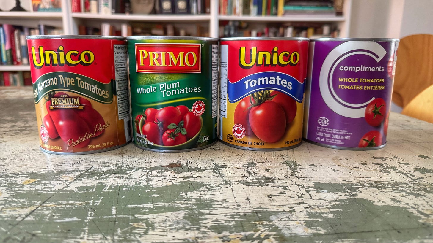 Cans of tomatoes on a dining room table. From left to right: Unico San Marzano Type Tomatoes, Primo Whole Plum Tomatoes, Unico Tomatoes, Compliments Whole Tomatoes