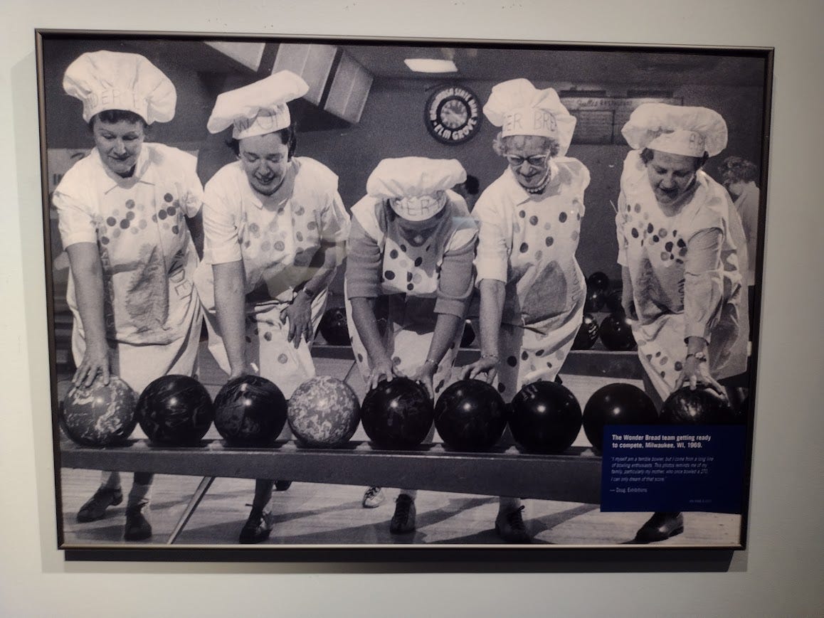 Women in puffy chef hats and spotted aprons in staged photo in bowling alley as they reach for bowling balls