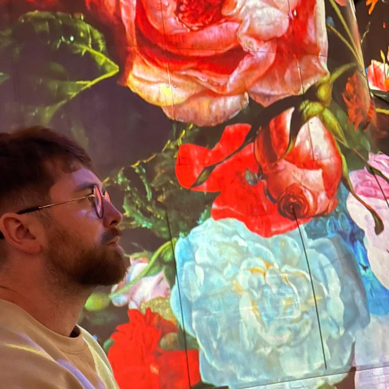 a man with circular, bronze-rimmed glasses looks at the projection of artwork on the walls around him in focused wonder
