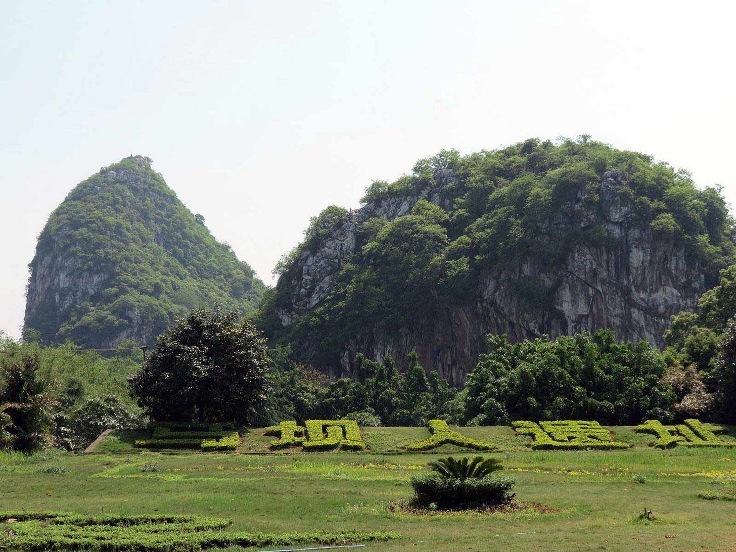 Two impressive hills with sheer rock cliffs bordering them. They are covered with green trees, and a hedge in the foreground is sculpted with Chinese characters Two impressive hills with sheer rock cliffs bordering them. They are covered with green trees, and a hedge in the foreground is sculpted with Chinese characters