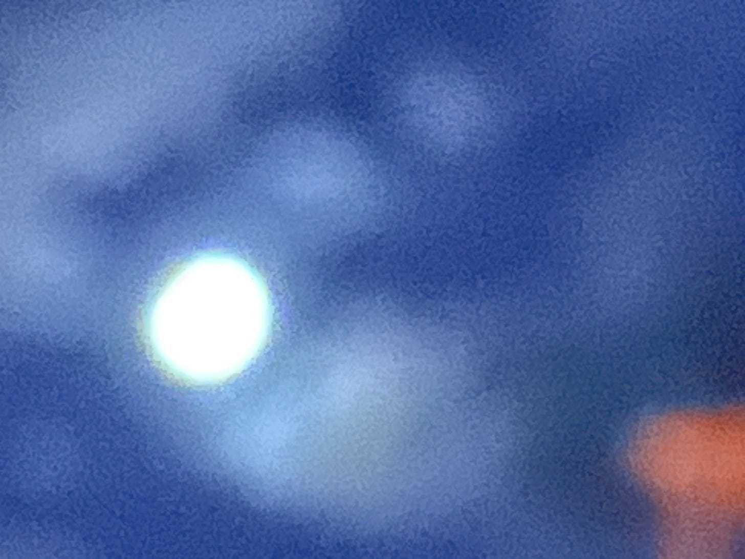 A very grainy but vividly blue photograph with a round white area, a moon or sun, offset to the left. An orange shape, perhaps the upper corner of a building is visible in the lower left.