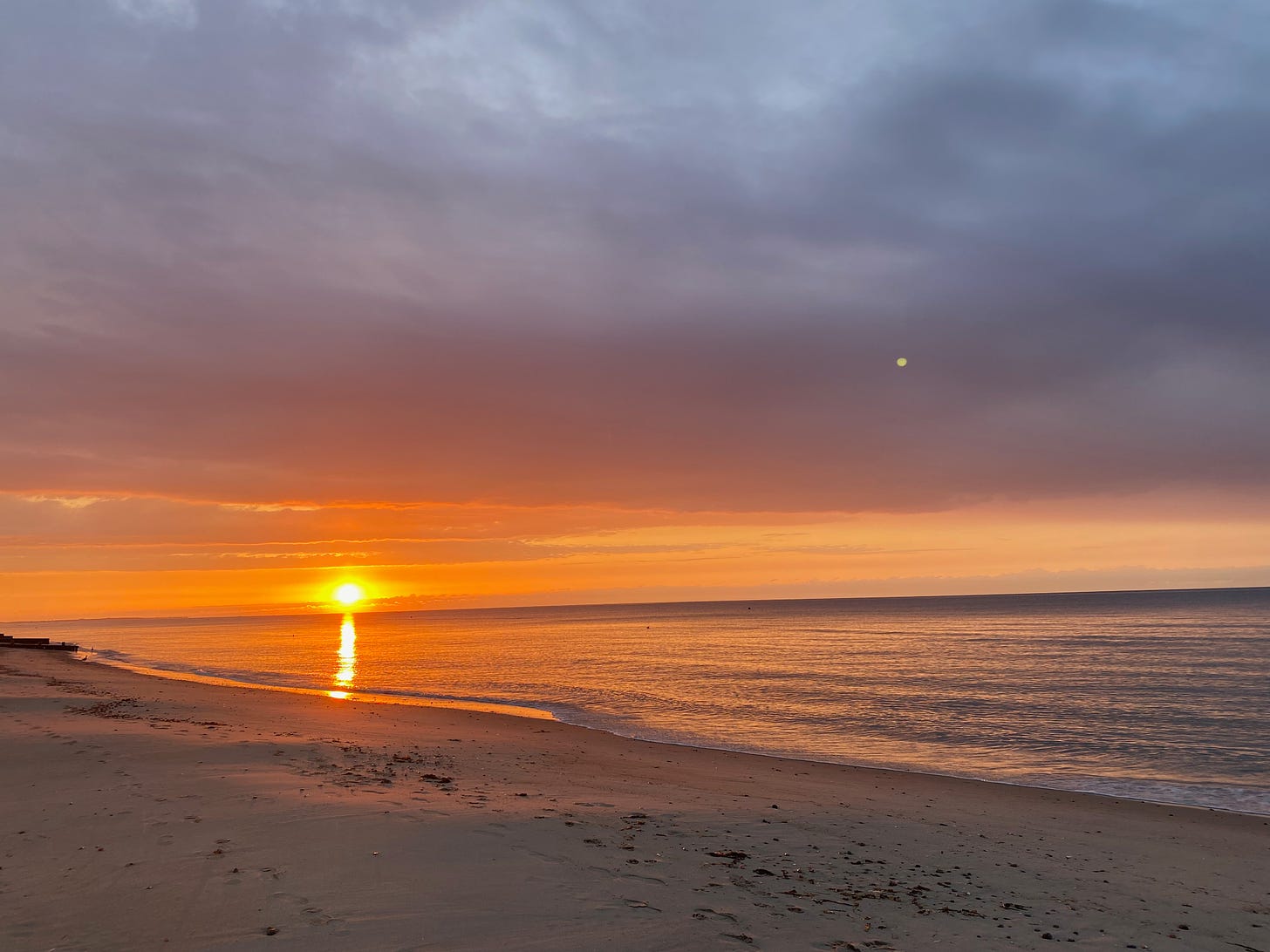 A beach at sunset. The sun leaves a brilliant, dazzling, golden trail across the water. The sky is orange and gold on the horizon, with pink and blue clouds above. The ocean is shimmering with golden light.