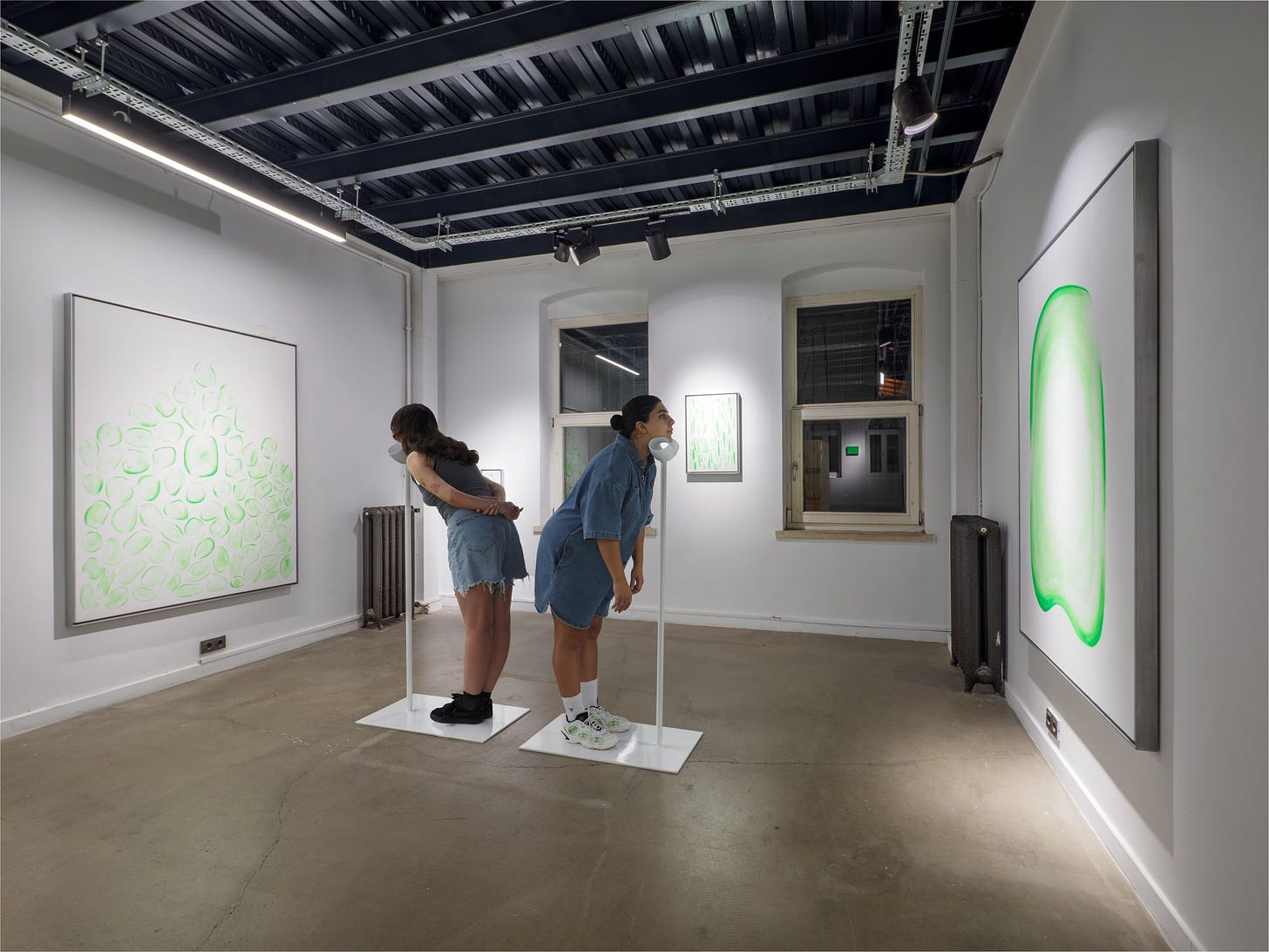 Two people stand and lean into thin white sticks which have chin cups. They appear to view green and white abstract art on the walls of a contemporary art gallery.