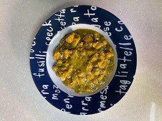 A khaki green bowl of curry. There are chunks of food amidst lots of sauce. In a bowl with a blue rim and white writing around the rim. The words are the names of pasta shapes.