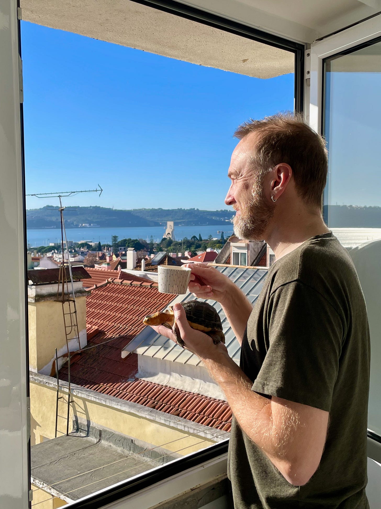 A man looks out at an open window at the blue sky over the rooftops of Lisbon, Portugal, while holding a cup of coffee in one hand and a turtle in the other.