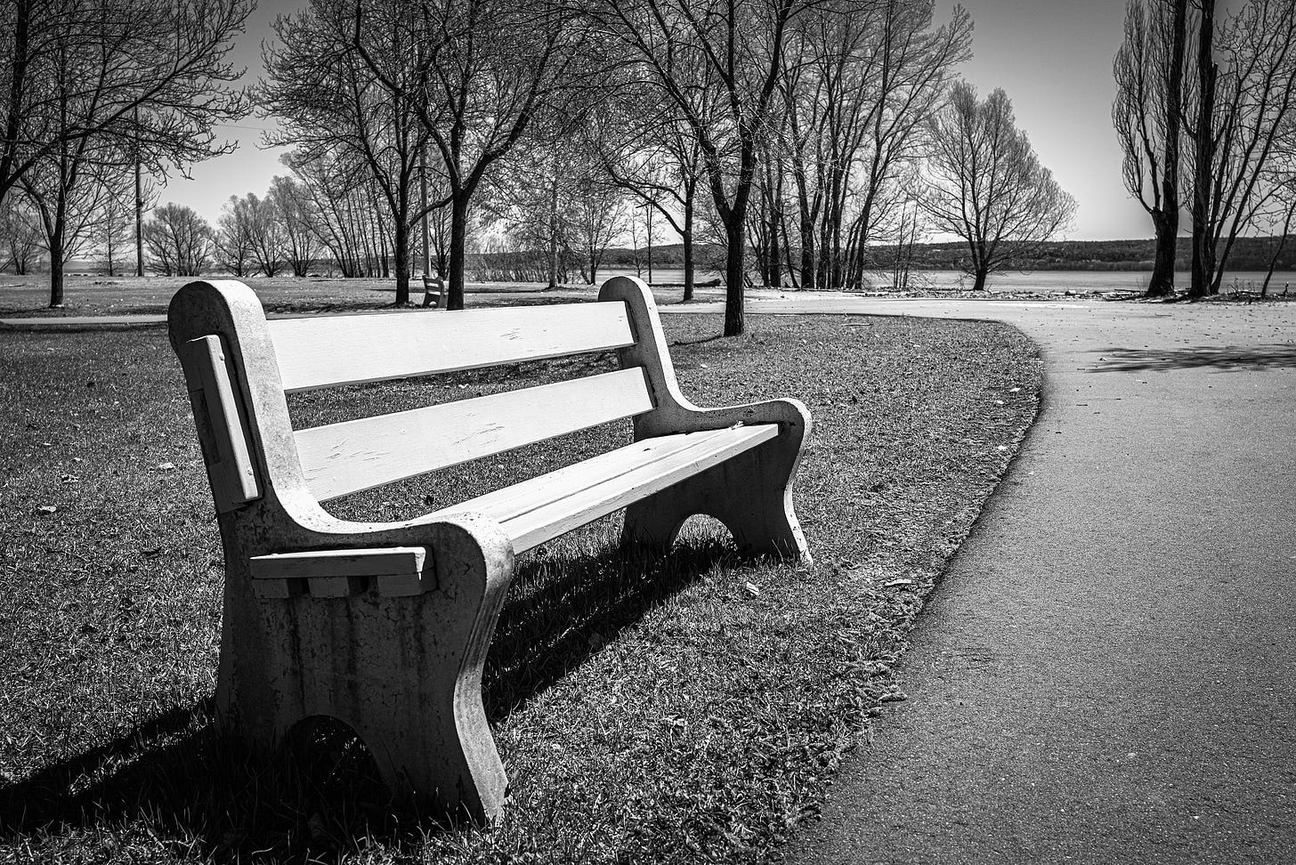 Black and white photograph of an empty park bench near a paved pathway in early spring