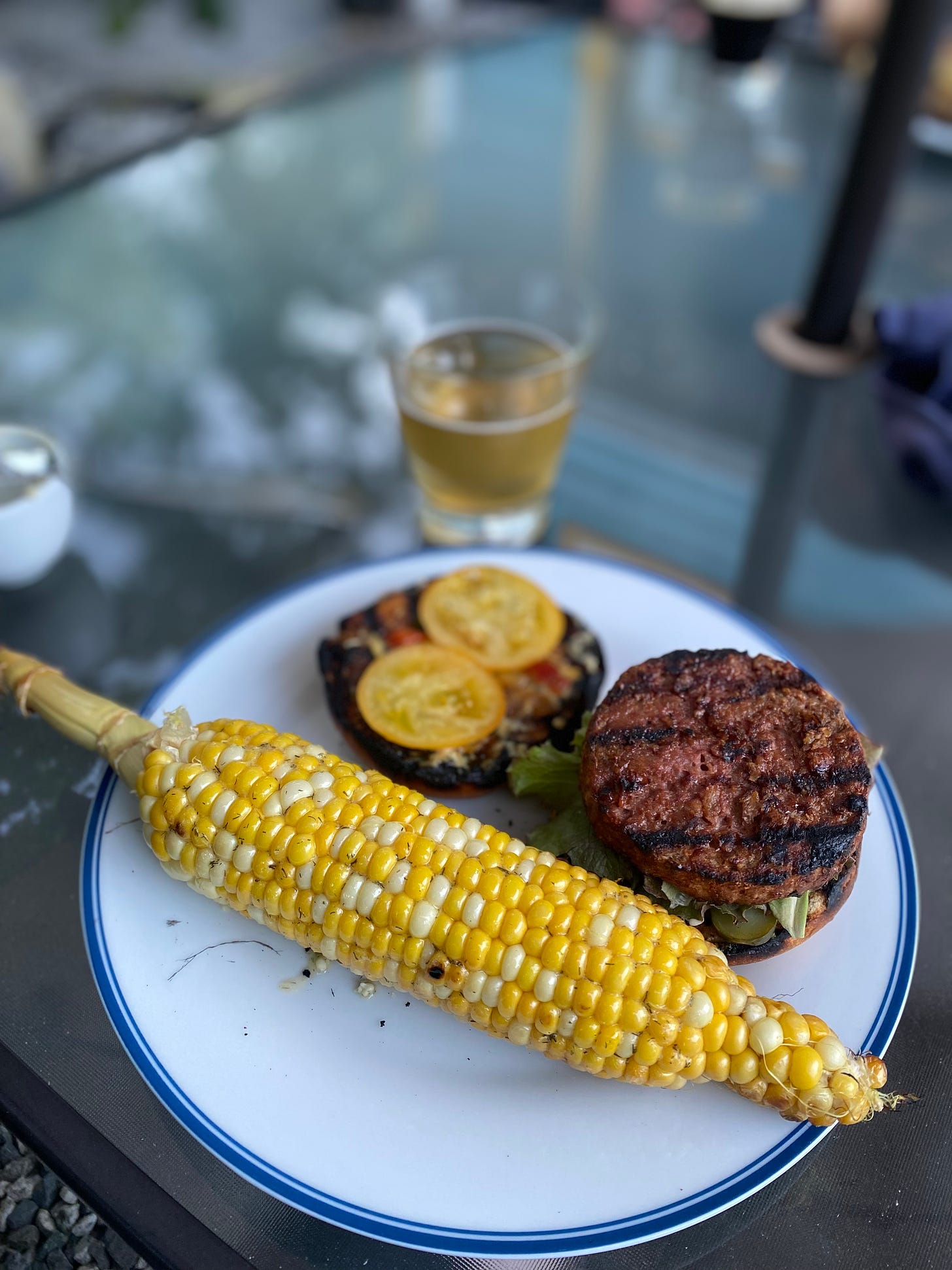 On a white plate, an ear of grilled corn with dill butter. Behind the corn is a burger on a grilled bun, still open to show a grilled Beyond patty on top of lettuce and pickle, the top half with two slices of yellow heirloom tomato. Behind the plate is a partially empty glass of light beer.