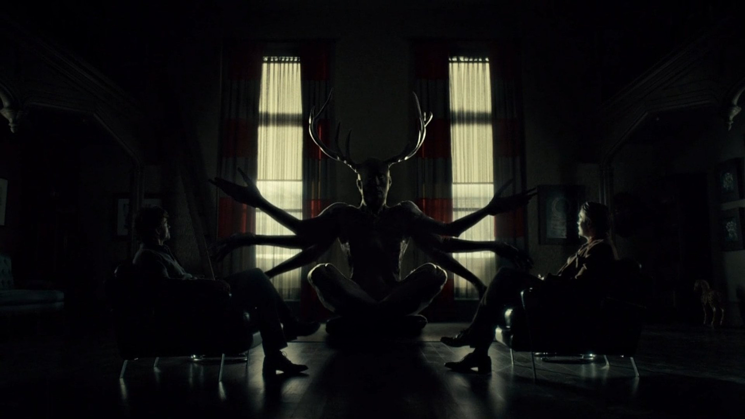 A screenshot from the television show Hannibal, showing Will and Hannibal sitting across from each other with a w*ndigo-like demon between them.