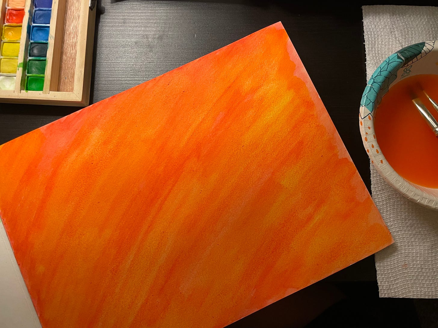 A paper painted over with water colors in orange, red, and yellow. Off to the sides are a palette of colors and a bowl full of painty water. 