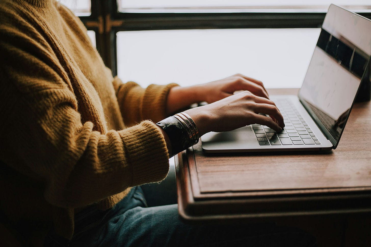 Side view of a person in a yellow sweater typing on a laptop.