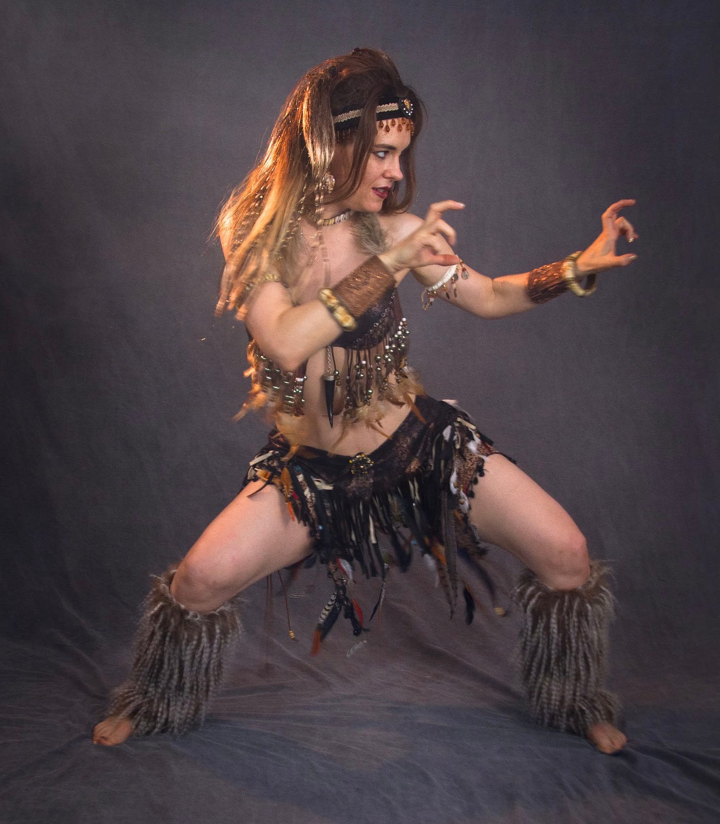 The author in a wild woman costume of fur, feathers, and fringe, in a deep stance with clawed hands at the ready and a gaze speared upon her target.