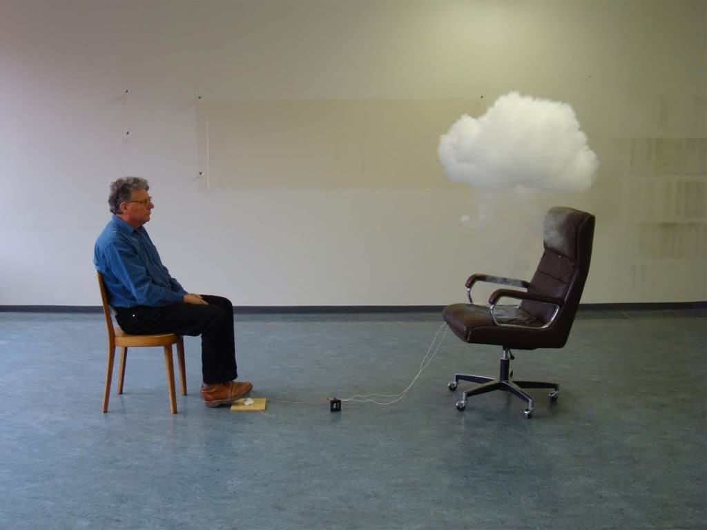 Artist Roman Signer, in black slacks and a blue shirt, sits in a wooden chair; across from him is a padded office chair connected to wires; a puff of smoke floats above the chair